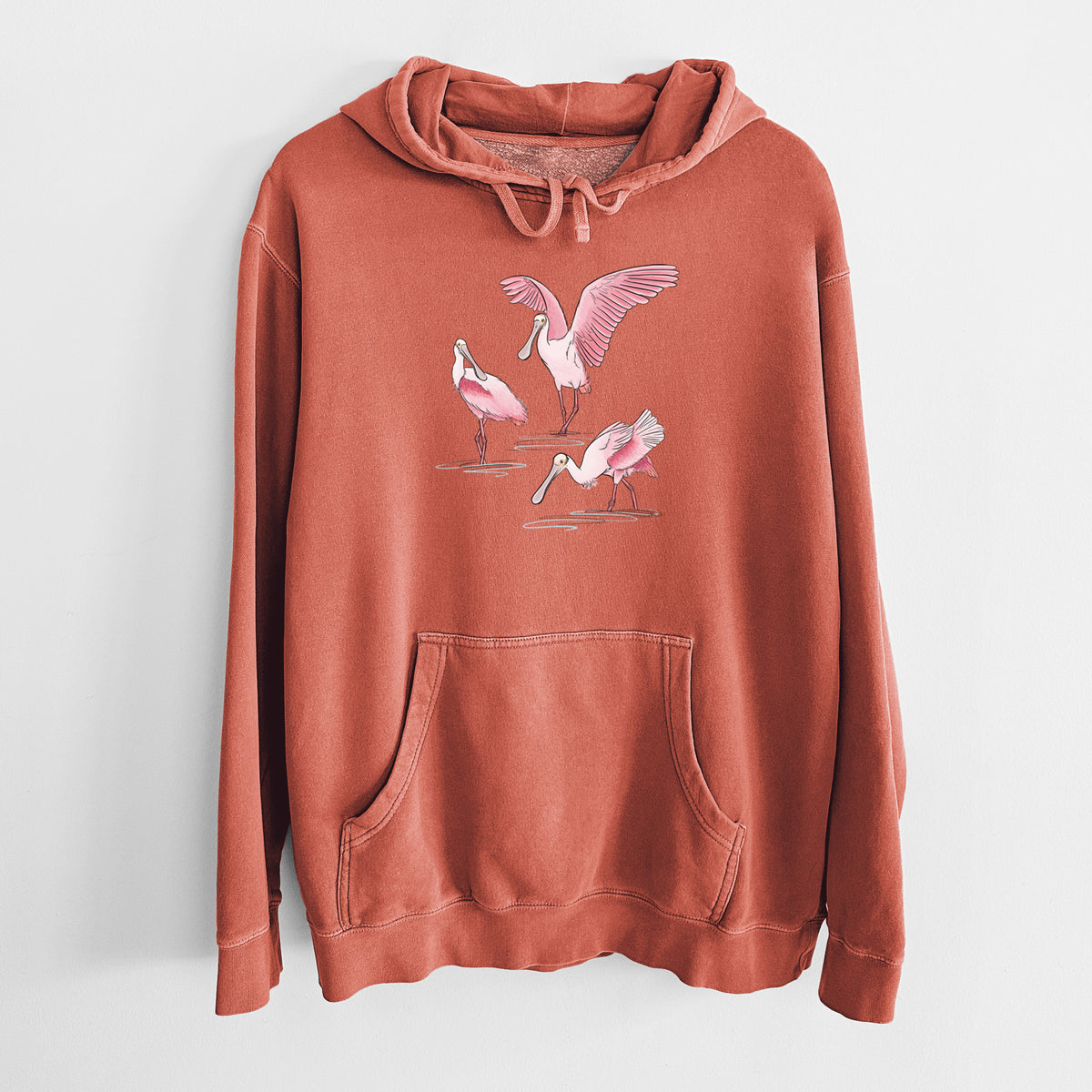 Amber orange pigment dyed hoodie laid flat, printed with a full-color illustration of three pink roseate spoonbills wading, one with its wings spread.
