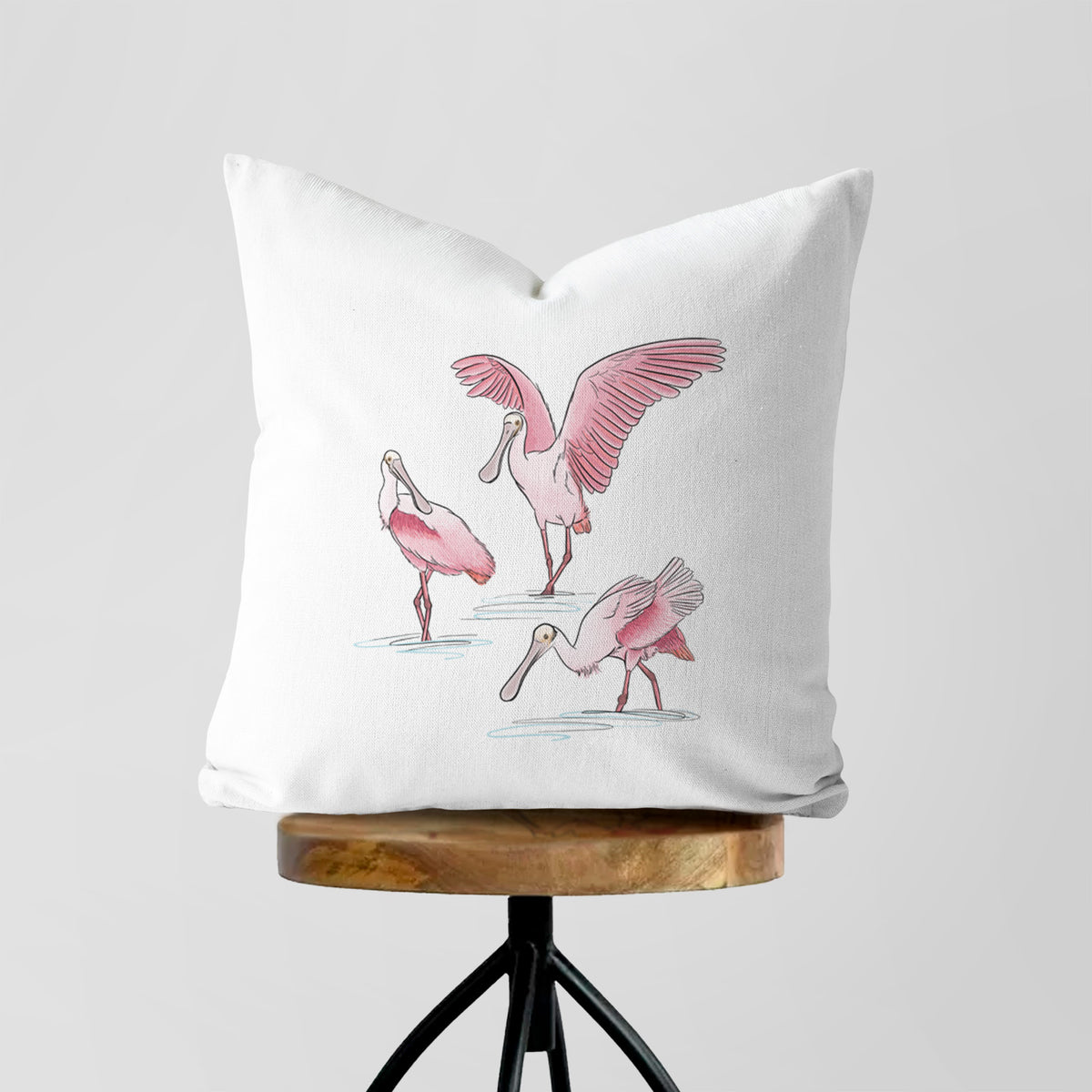 White pillow cover printed with a full-color illustration of three pink roseate spoonbills wading, one with its wings spread, on a wooden stool.