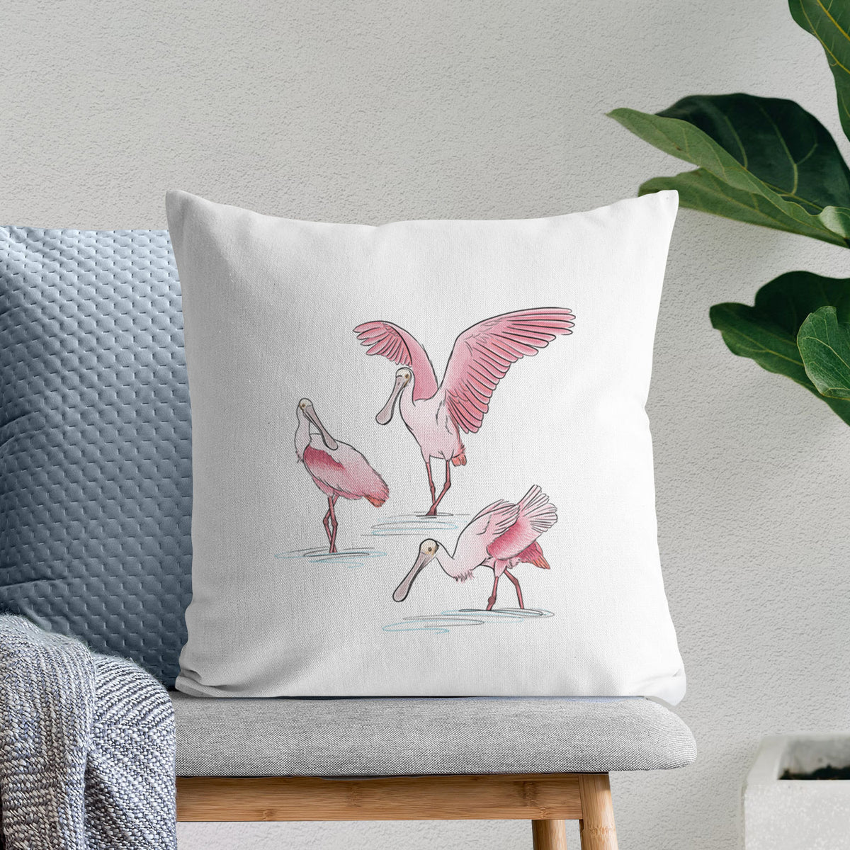 White pillow cover printed with a full-color illustration of three pink roseate spoonbills wading, one with its wings spread, on a bench beside plants.