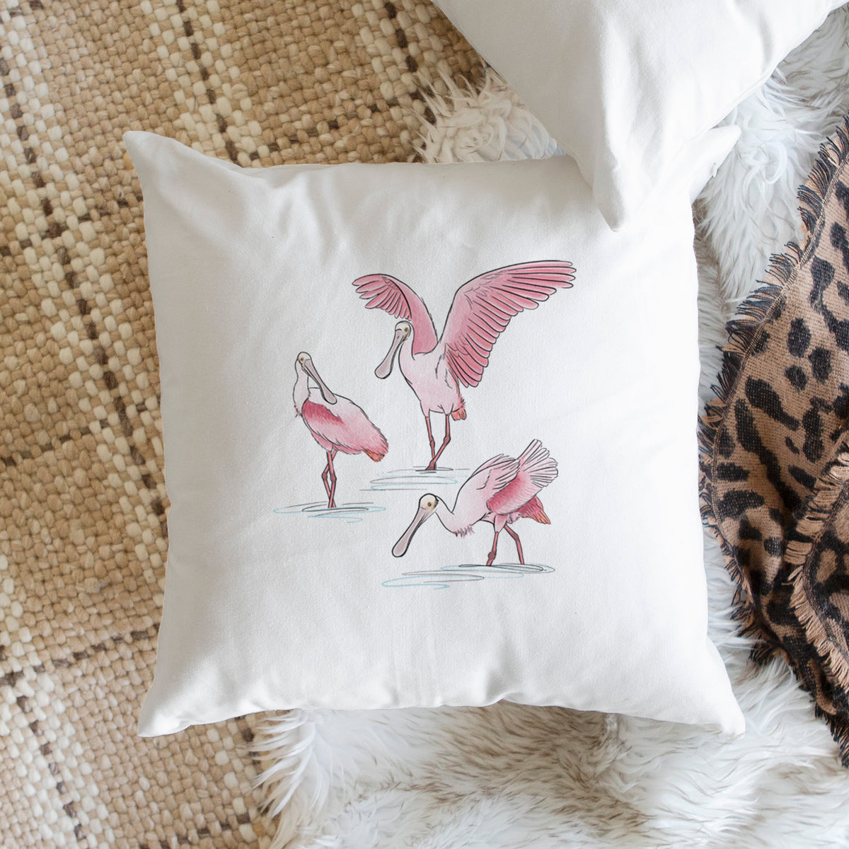 White pillow cover printed with a full-color illustration of three pink roseate spoonbills wading, one with its wings spread, on a woven chair.