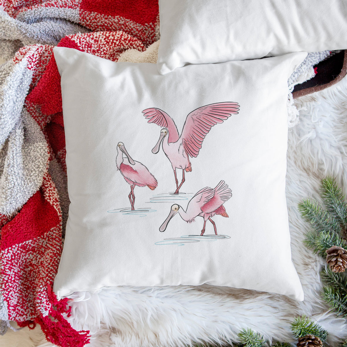 White pillow cover printed with a full-color illustration of three pink roseate spoonbills wading, one with its wings spread, on a woven chair.