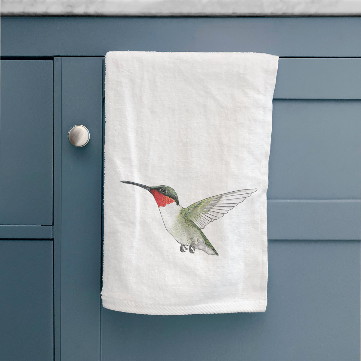 White hand towel printed with a full-color illustration of a ruby-throated hummingbird in flight, hanging on a cabinet door.