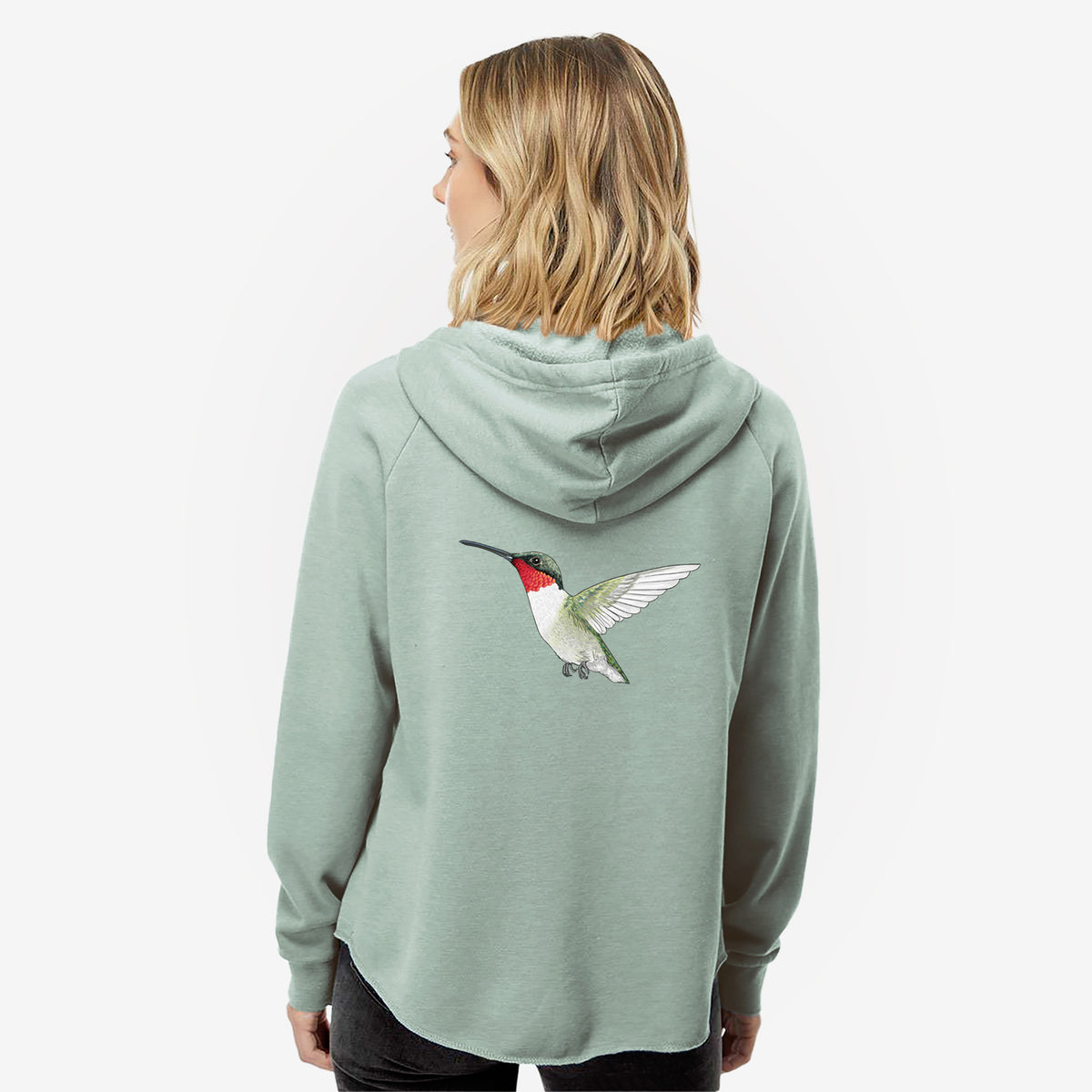 Woman wearing a sage green women's Cali Wave zip-up sweatshirt printed with a full-color illustration of a ruby-throated hummingbird in flight, seen from behind.