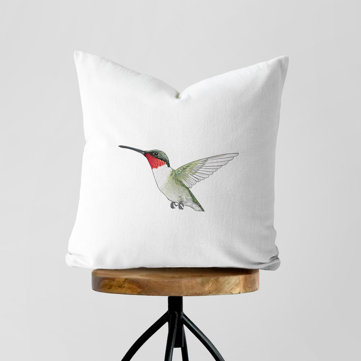 White pillow cover printed with a full-color illustration of a ruby-throated hummingbird in flight, on a wooden stool.