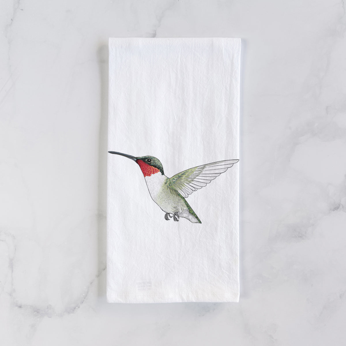 White cotton tea towel laid flat on marble, printed with a full-color illustration of a ruby-throated hummingbird in flight.