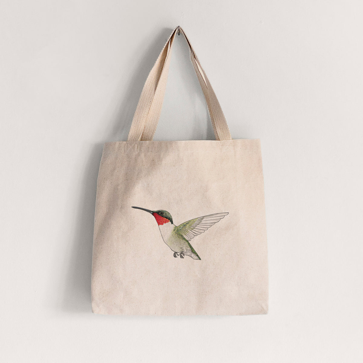 Natural canvas tote bag printed with a full-color illustration of a ruby-throated hummingbird in flight.