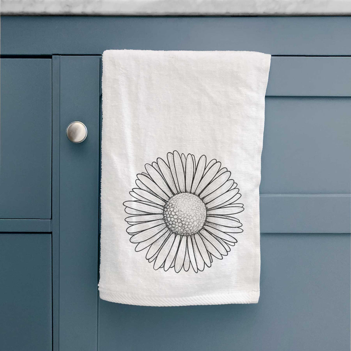 White hand towel printed with a detailed black ink line drawing of a single common daisy flower, hanging on a cabinet door.