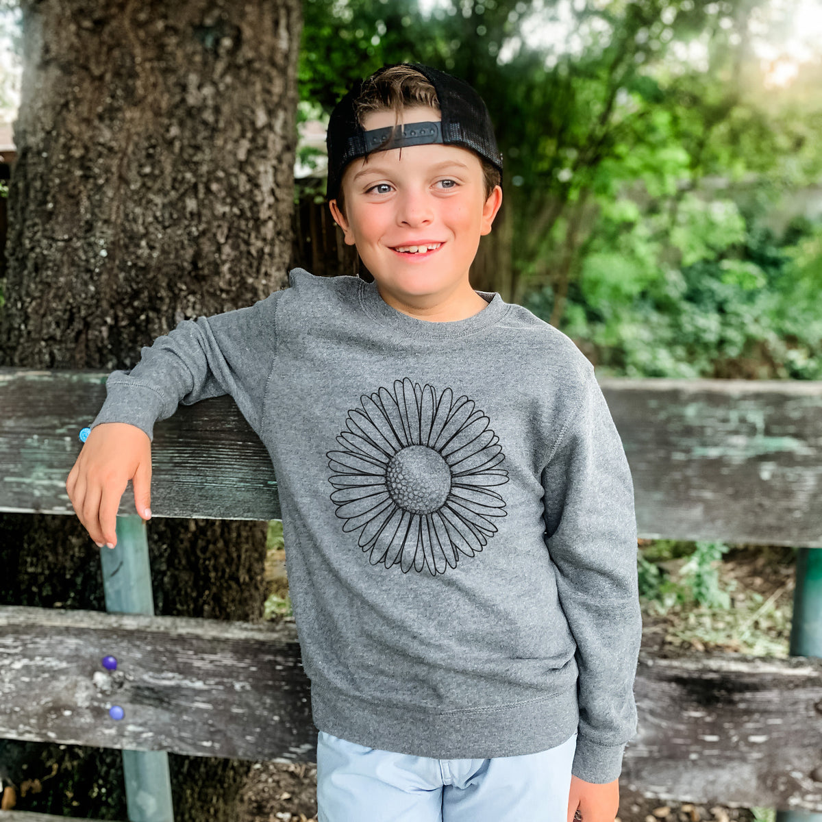 Boy wearing a youth crewneck sweatshirt printed with a detailed black ink line drawing of a single common daisy flower, outdoors.