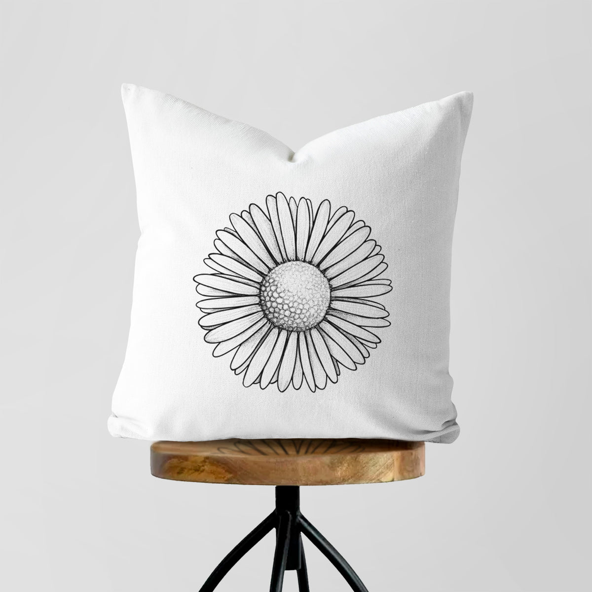 White pillow cover printed with a detailed black ink line drawing of a single common daisy flower, on a wooden stool.