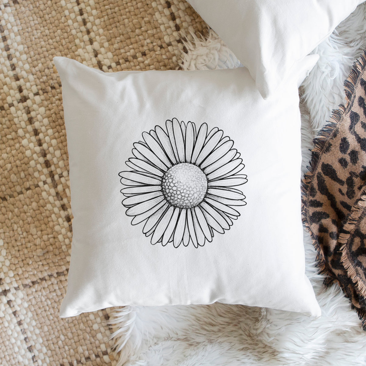 White pillow cover printed with a detailed black ink line drawing of a single common daisy flower, on a woven chair.