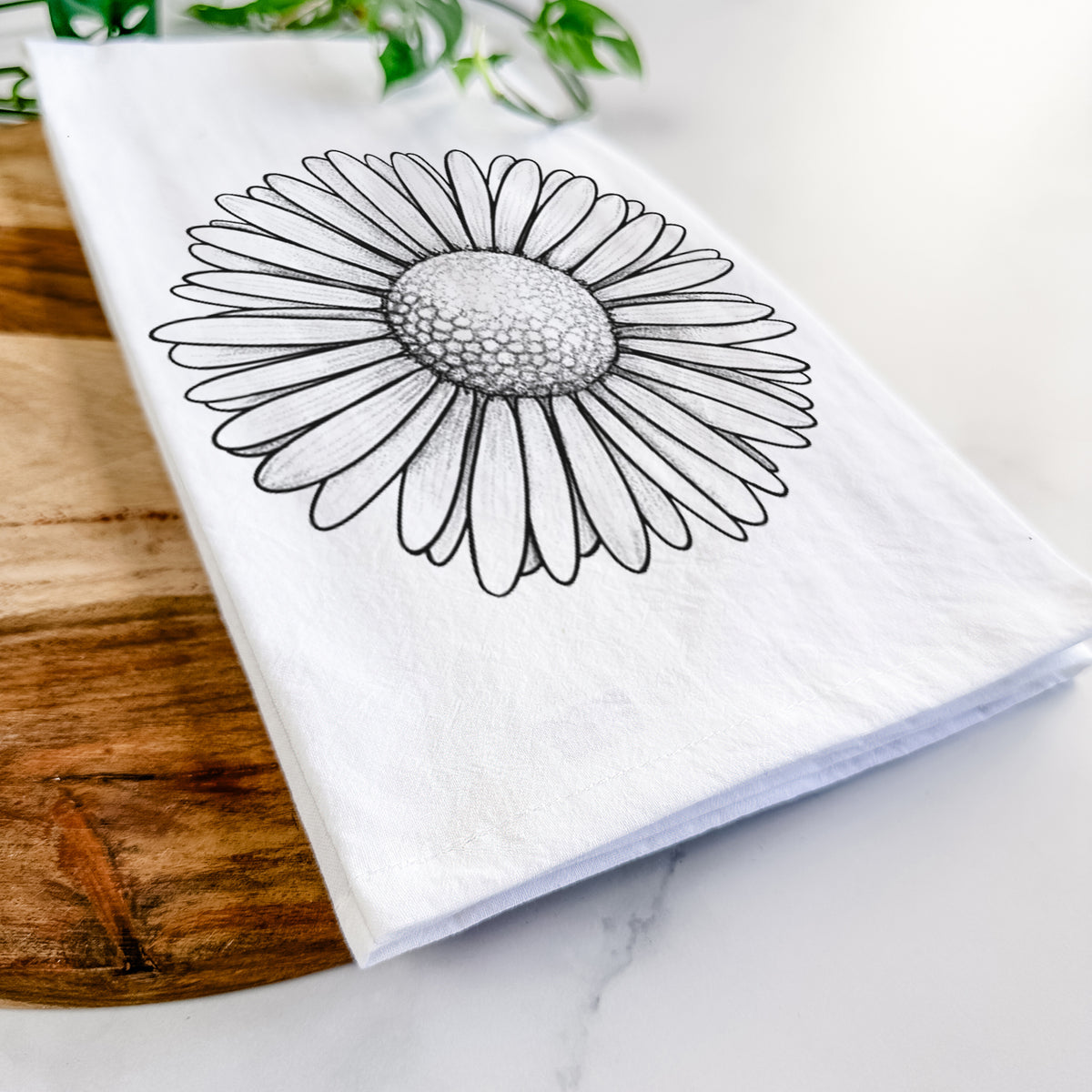 Close-up of a folded white tea towel on a wooden board, showing a detailed black ink line drawing of a single common daisy flower.