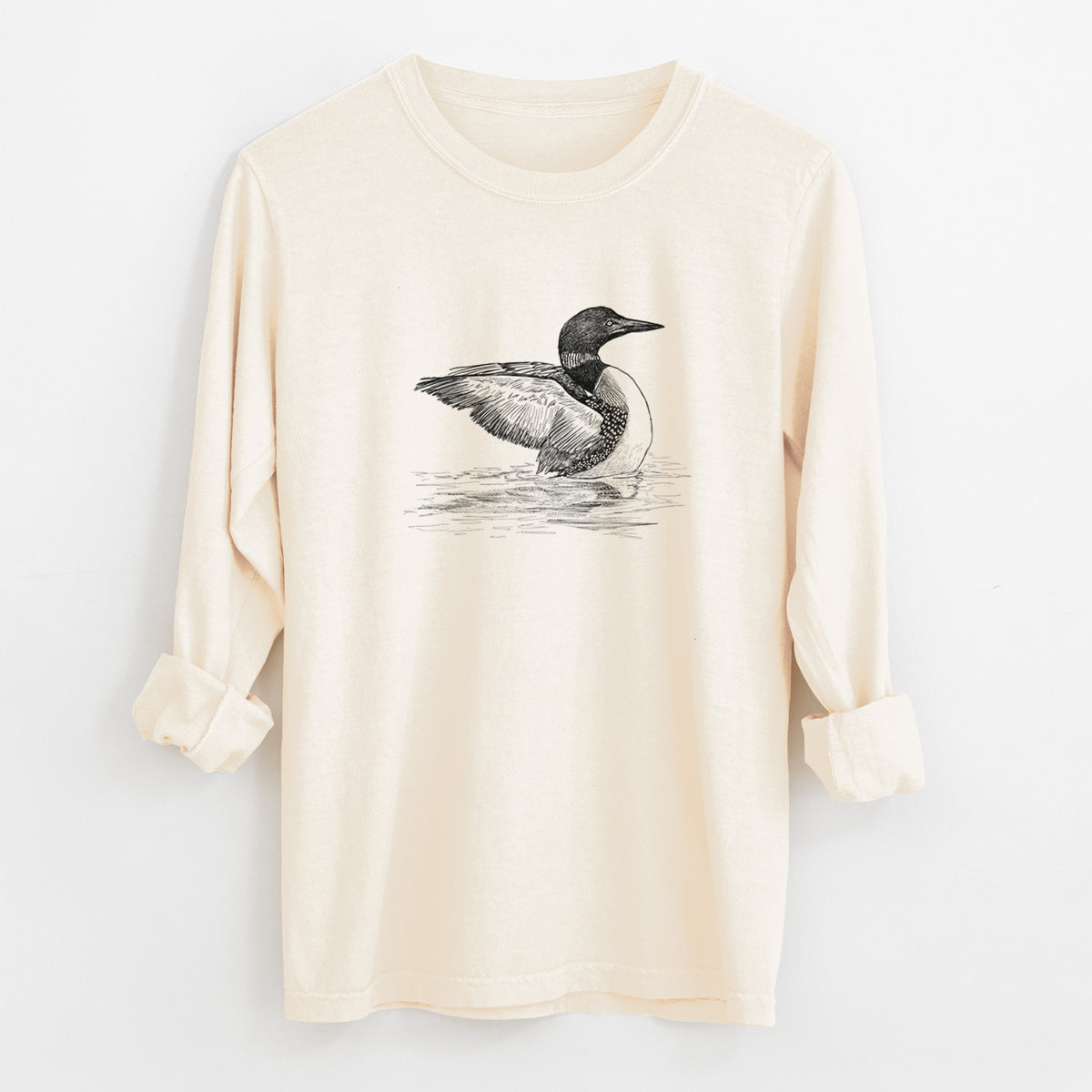 Ivory Comfort Colors long sleeve shirt laid flat with the cuffs rolled, printed with a detailed black ink sketch of a common loon floating on rippling water.