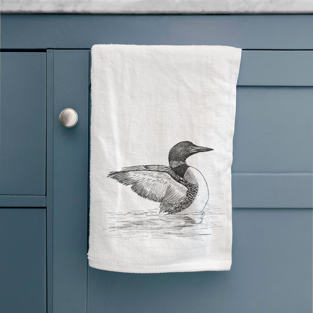 White hand towel printed with a detailed black ink sketch of a common loon floating on rippling water, hanging on a cabinet door.