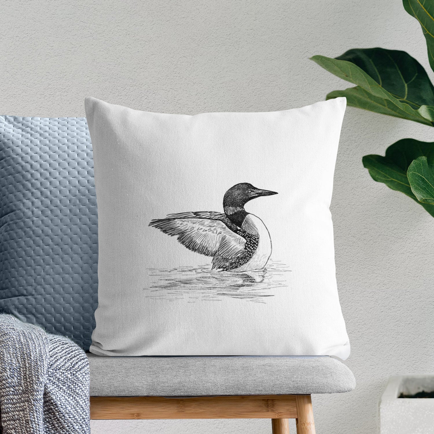 White pillow cover printed with a detailed black ink sketch of a common loon floating on rippling water, on a bench beside plants.