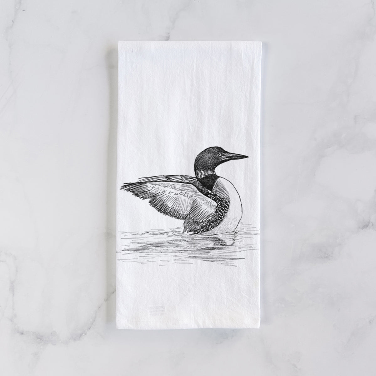 White cotton tea towel laid flat on marble, printed with a detailed black ink sketch of a common loon floating on rippling water.