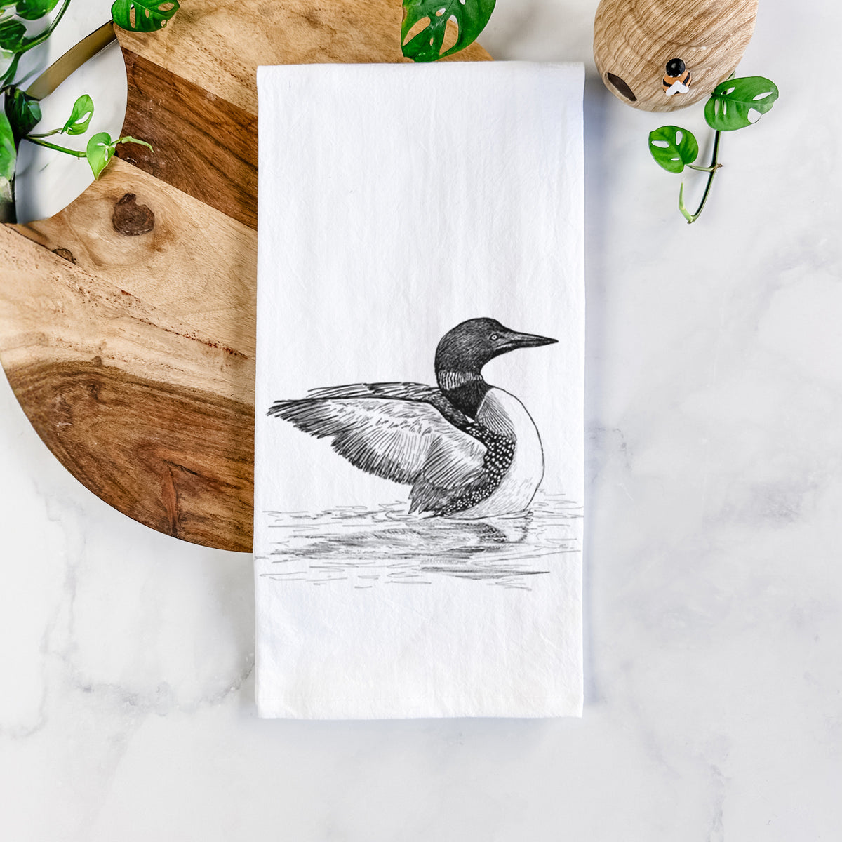White cotton tea towel printed with a detailed black ink sketch of a common loon floating on rippling water, on a marble counter beside a round wooden cutting board and greenery.