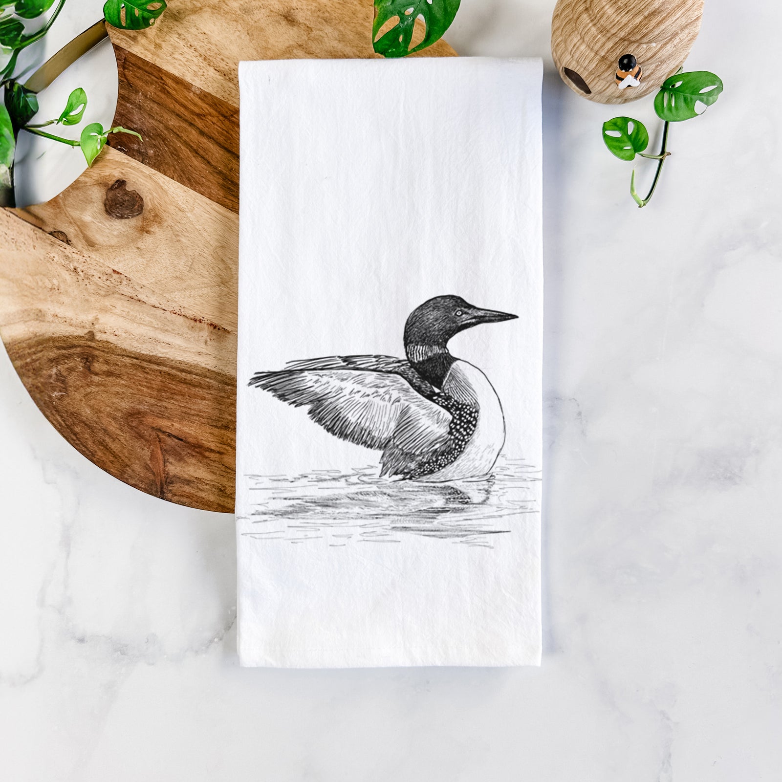 White cotton tea towel printed with a detailed black ink sketch of a common loon floating on rippling water, on a marble counter beside a round wooden cutting board and greenery.