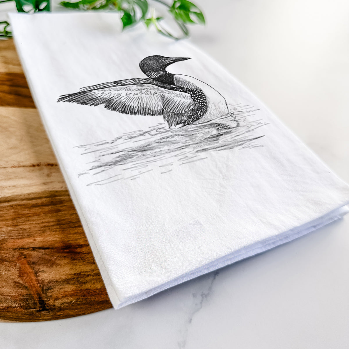 Close-up of a folded white tea towel on a wooden board, showing a detailed black ink sketch of a common loon floating on rippling water.