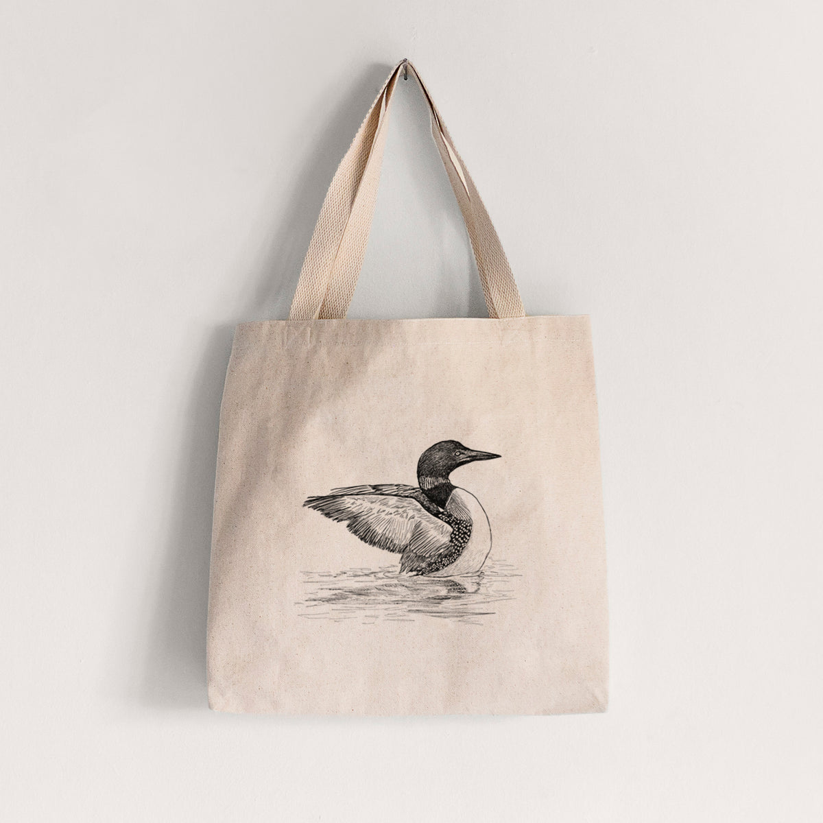 Natural canvas tote bag printed with a detailed black ink sketch of a common loon floating on rippling water.