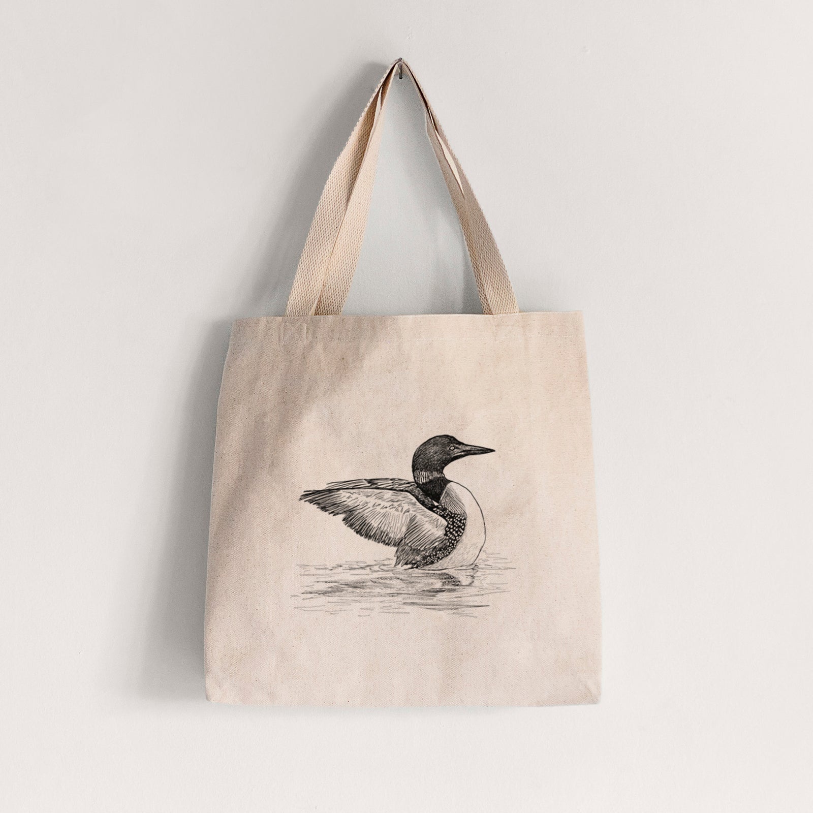 Natural canvas tote bag printed with a detailed black ink sketch of a common loon floating on rippling water.