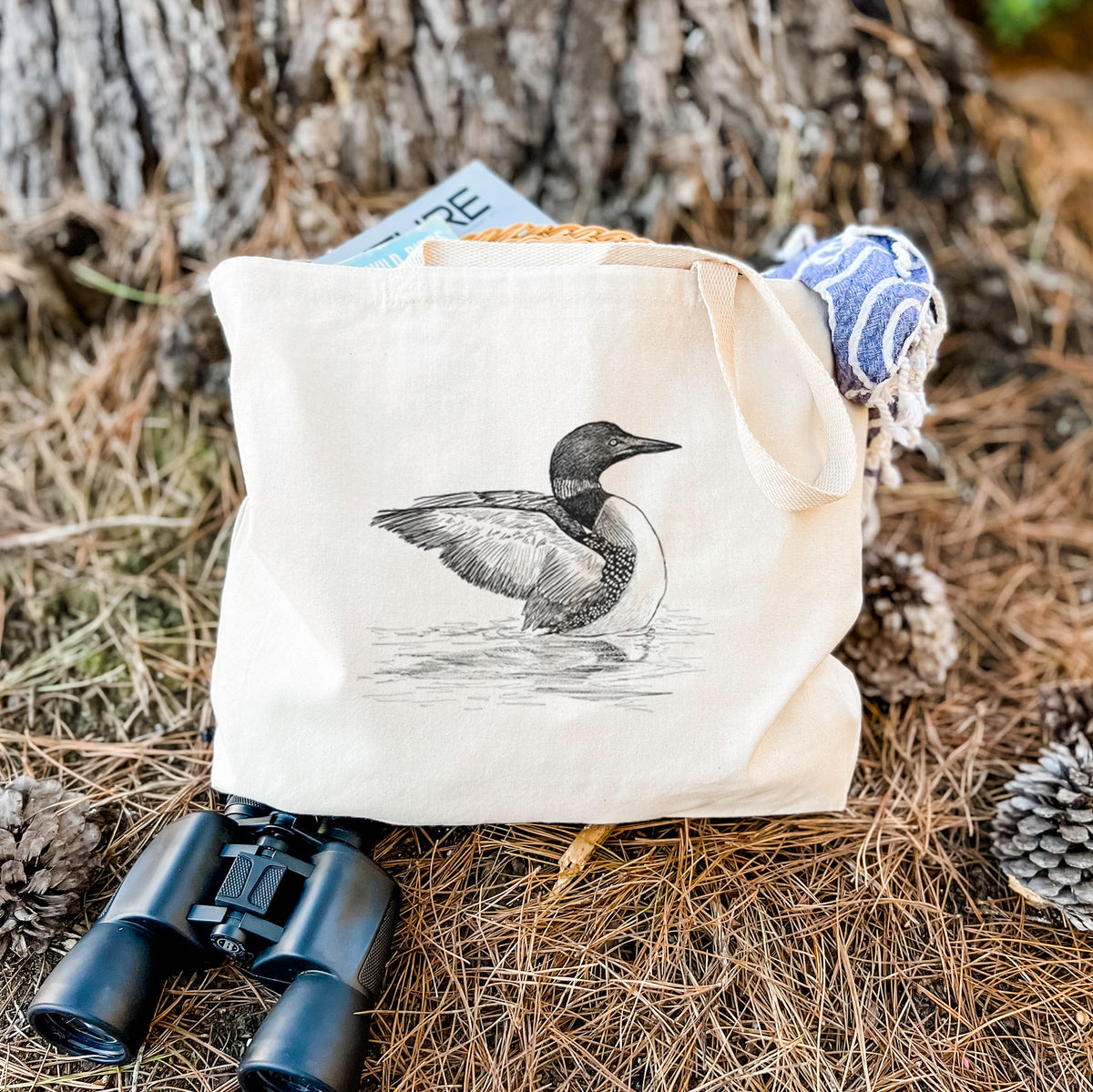 Canvas tote bag printed with a detailed black ink sketch of a common loon floating on rippling water, resting on dry grass beside a pair of binoculars.