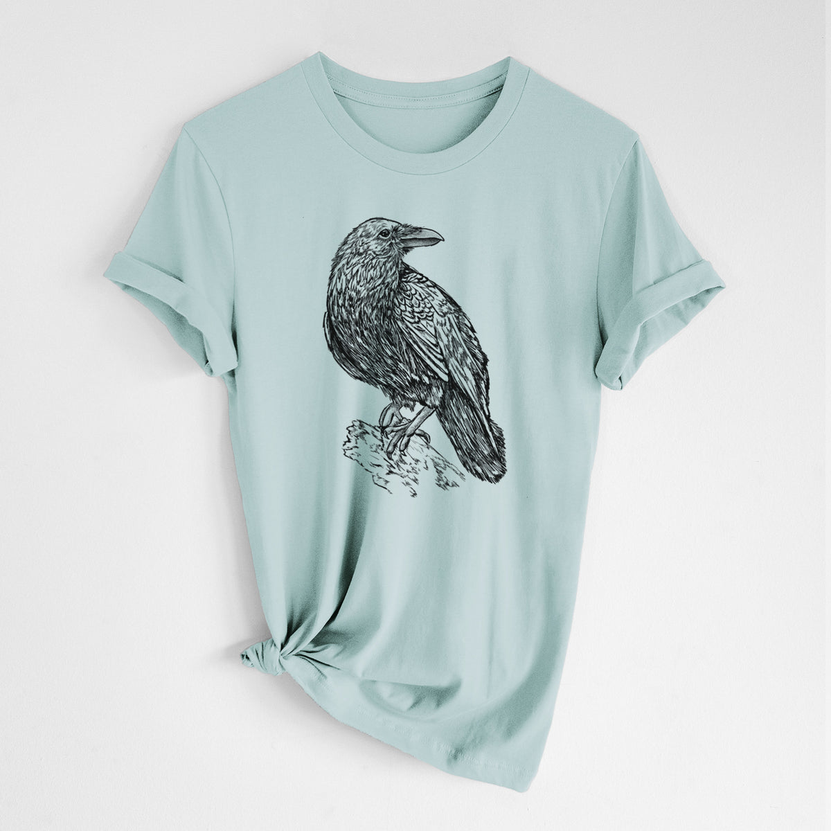 Dusty blue lightweight cotton t-shirt laid flat with the sleeves rolled, printed with a detailed black ink sketch of a common raven perched on a rock.