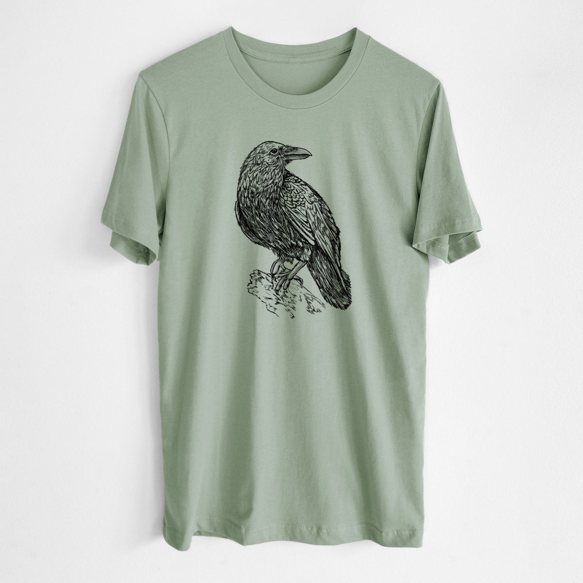 Sage green lightweight cotton t-shirt laid flat, printed with a detailed black ink sketch of a common raven perched on a rock.