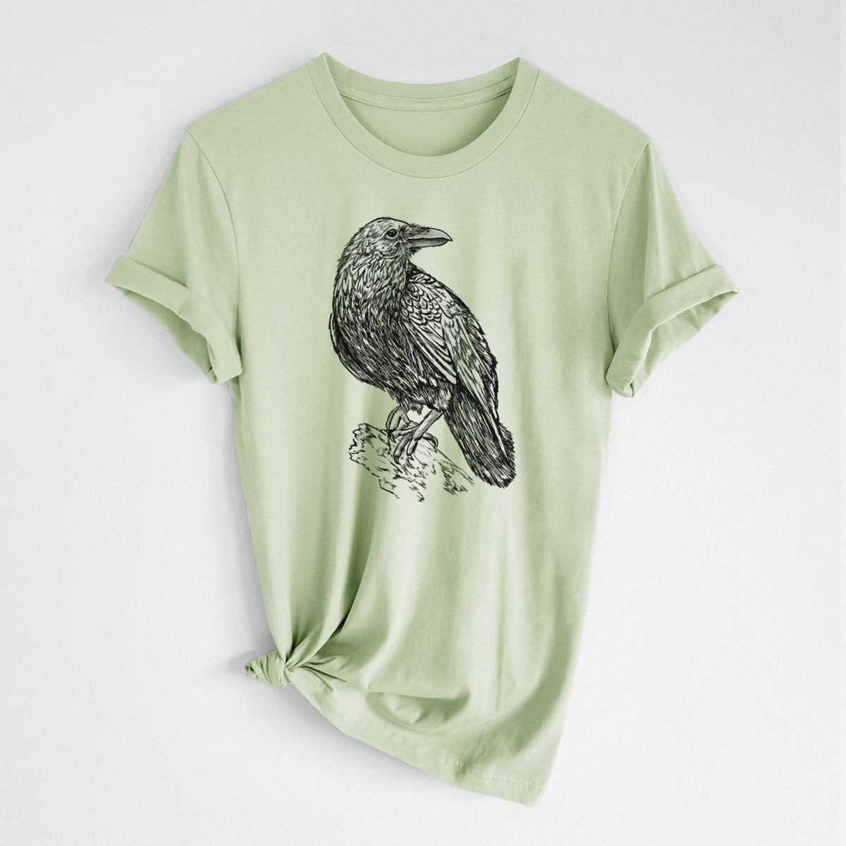 Spring green lightweight cotton t-shirt laid flat with the sleeves rolled, printed with a detailed black ink sketch of a common raven perched on a rock.