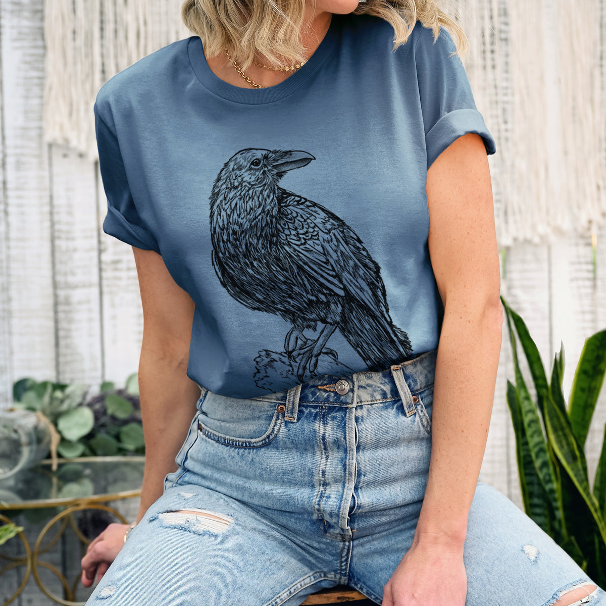 Woman wearing a steel blue lightweight cotton t-shirt printed with a detailed black ink sketch of a common raven perched on a rock.