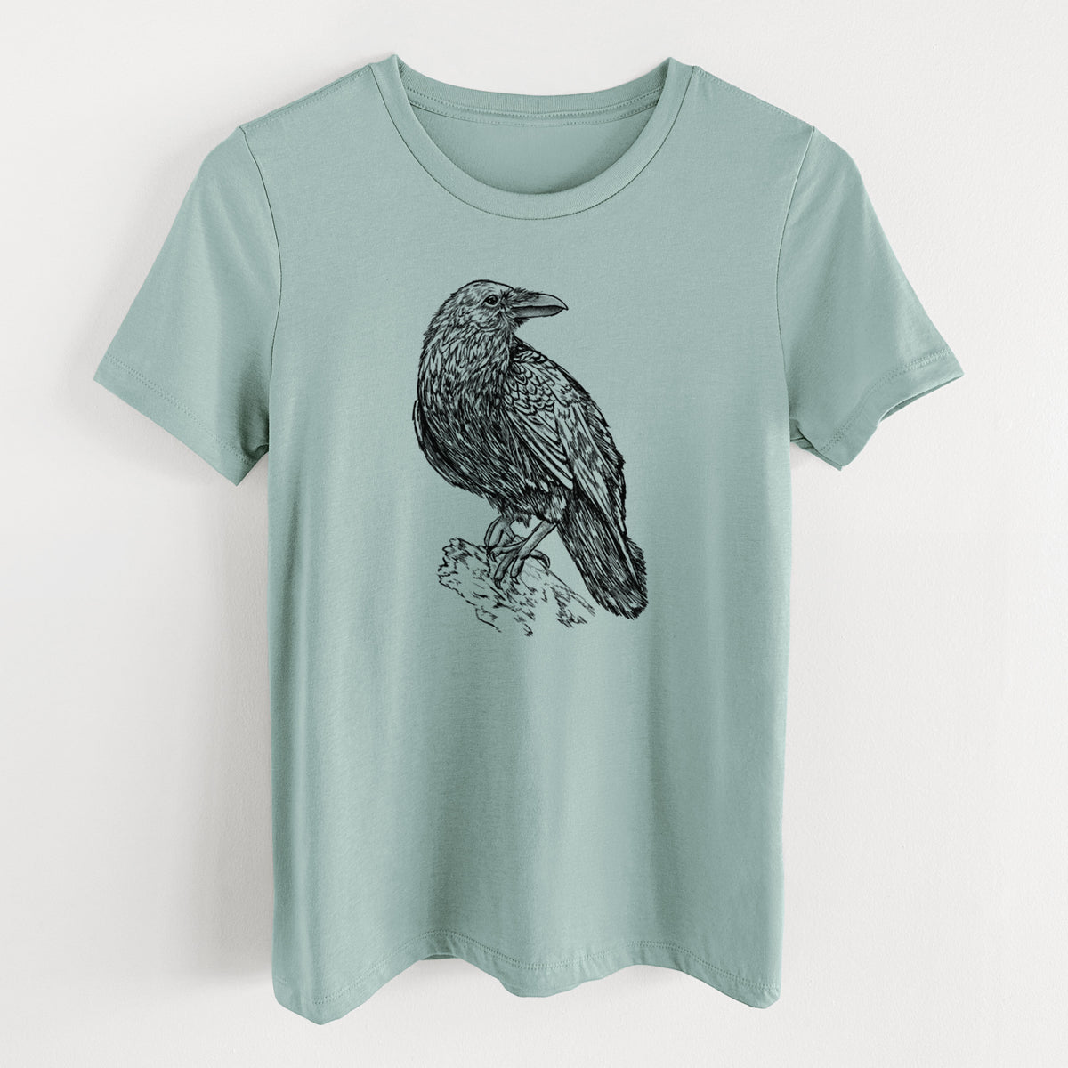 Dusty blue women's relaxed fit cotton t-shirt laid flat, printed with a detailed black ink sketch of a common raven perched on a rock.