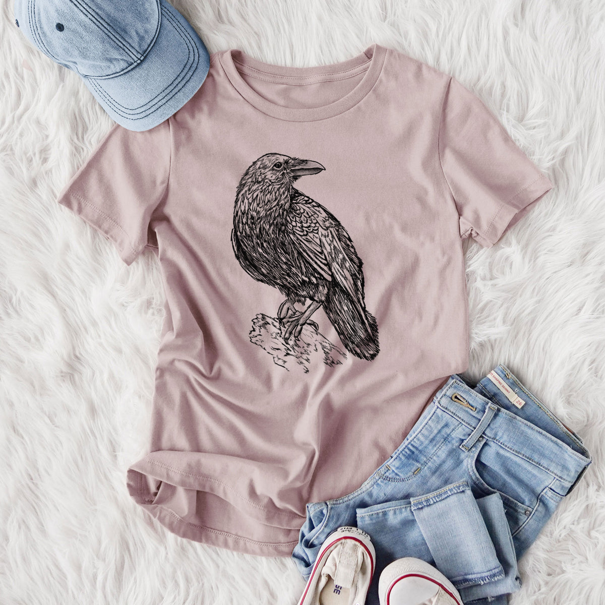 Women's relaxed fit cotton t-shirt printed with a detailed black ink sketch of a common raven perched on a rock, styled flat on a white faux fur rug with jeans.