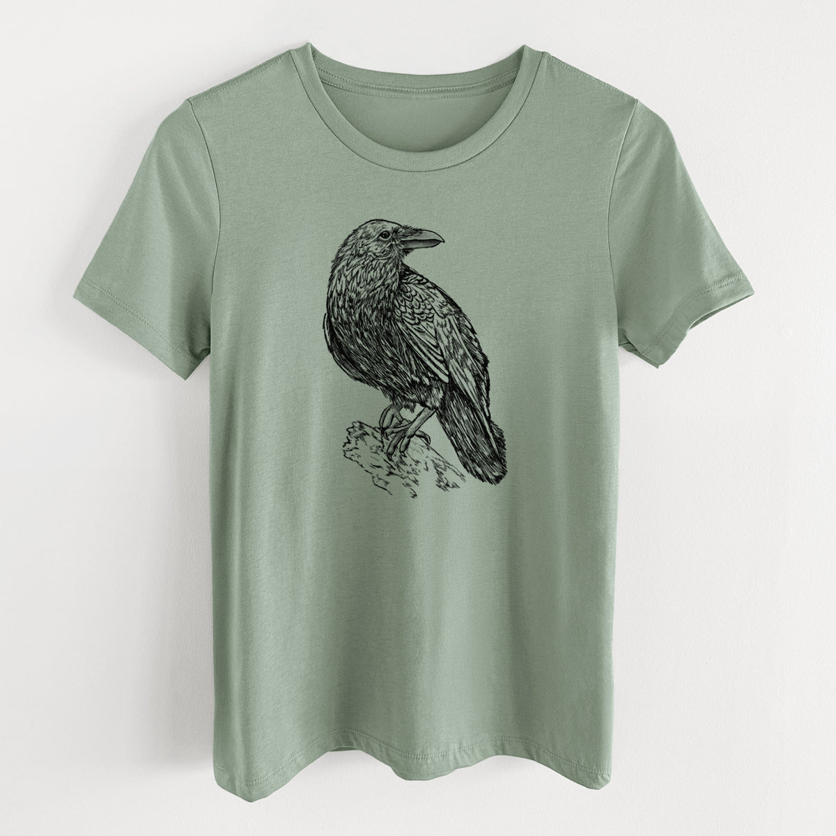 Sage green women's relaxed fit cotton t-shirt laid flat, printed with a detailed black ink sketch of a common raven perched on a rock.