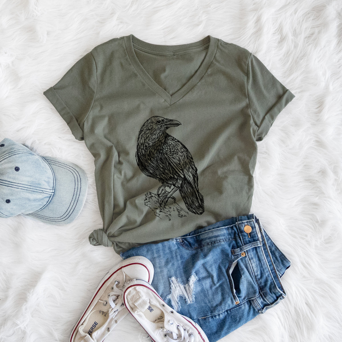 Military green women's relaxed fit cotton V-neck t-shirt printed with a detailed black ink sketch of a common raven perched on a rock, styled flat on a white faux fur rug with a cap, jeans and sneakers.