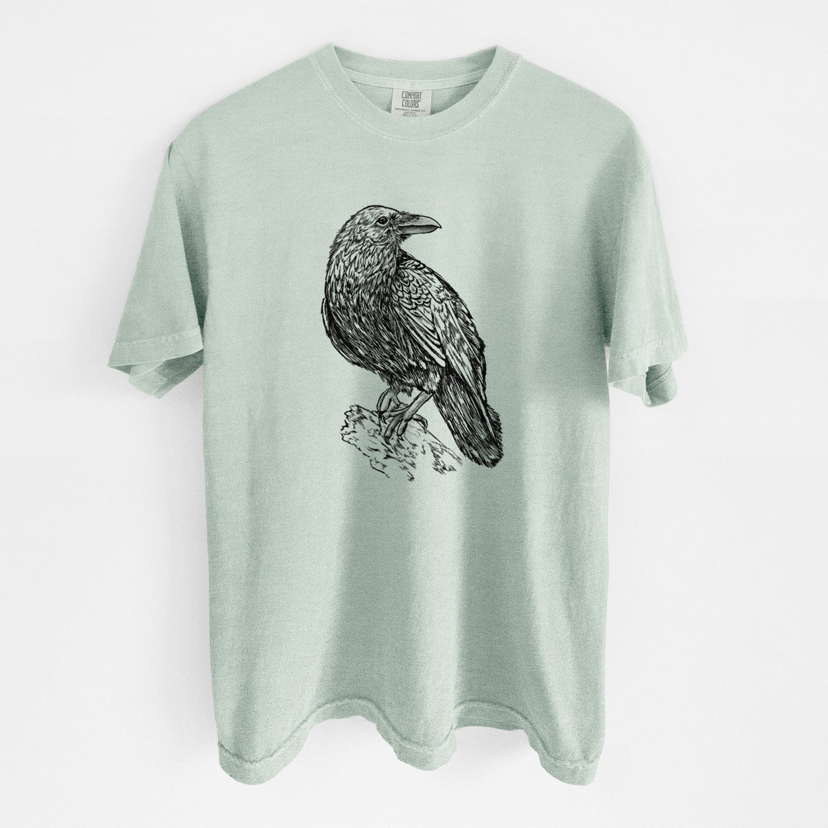 Bay green Comfort Colors t-shirt laid flat, printed with a detailed black ink sketch of a common raven perched on a rock.