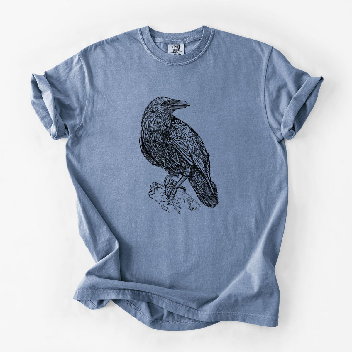 Blue jean Comfort Colors t-shirt laid flat, printed with a detailed black ink sketch of a common raven perched on a rock.