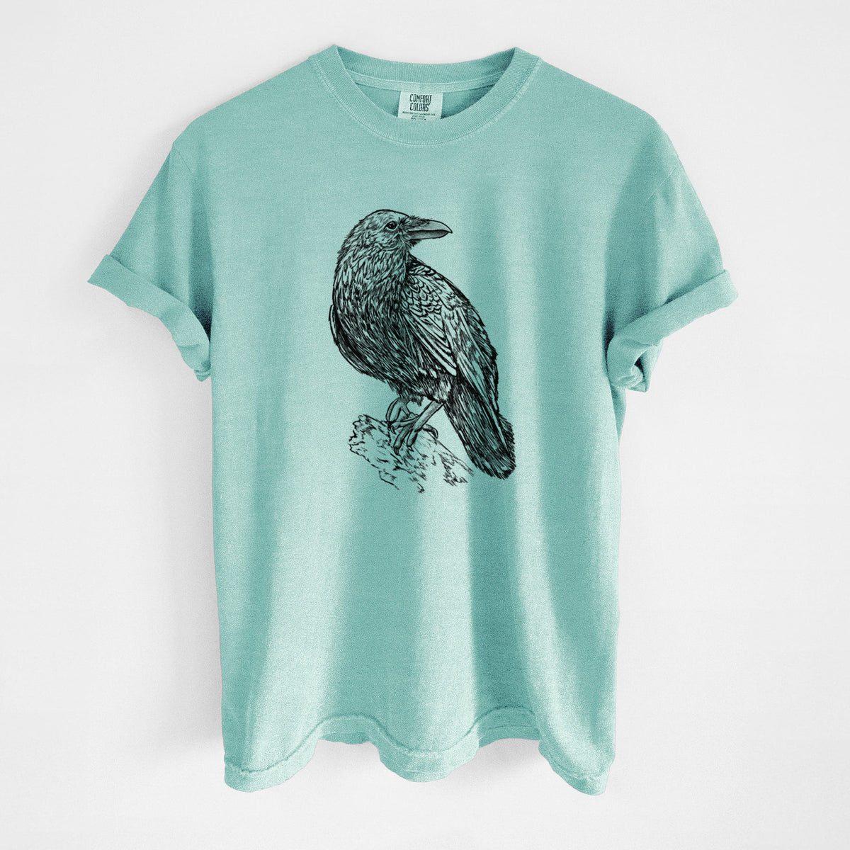 Chalky mint Comfort Colors t-shirt laid flat, printed with a detailed black ink sketch of a common raven perched on a rock.