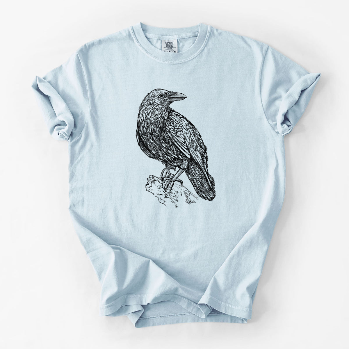 Chambray blue Comfort Colors t-shirt laid flat, printed with a detailed black ink sketch of a common raven perched on a rock.