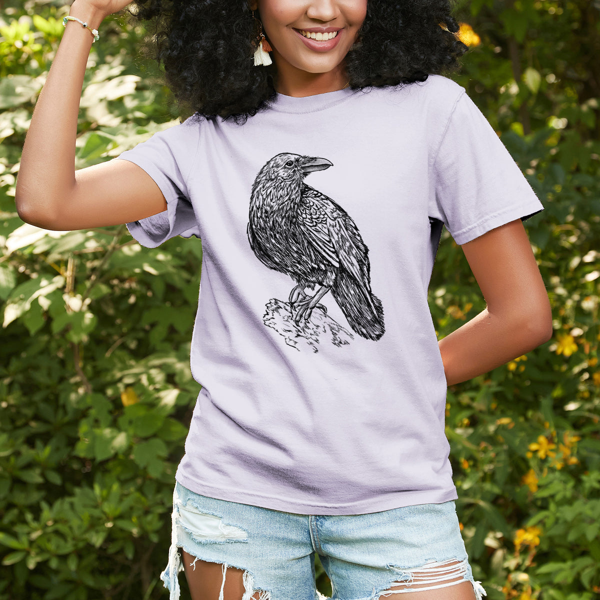 Woman wearing a Comfort Colors t-shirt printed with a detailed black ink sketch of a common raven perched on a rock, outdoors.