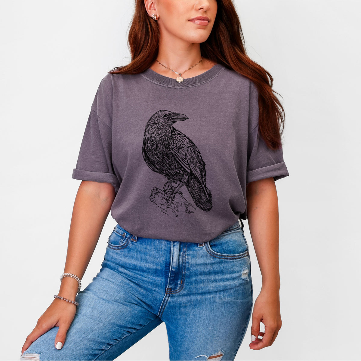 Woman wearing a Comfort Colors t-shirt printed with a detailed black ink sketch of a common raven perched on a rock.