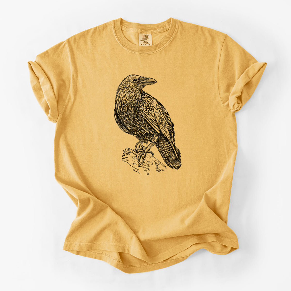 Golden wheat yellow Comfort Colors t-shirt laid flat, printed with a detailed black ink sketch of a common raven perched on a rock.