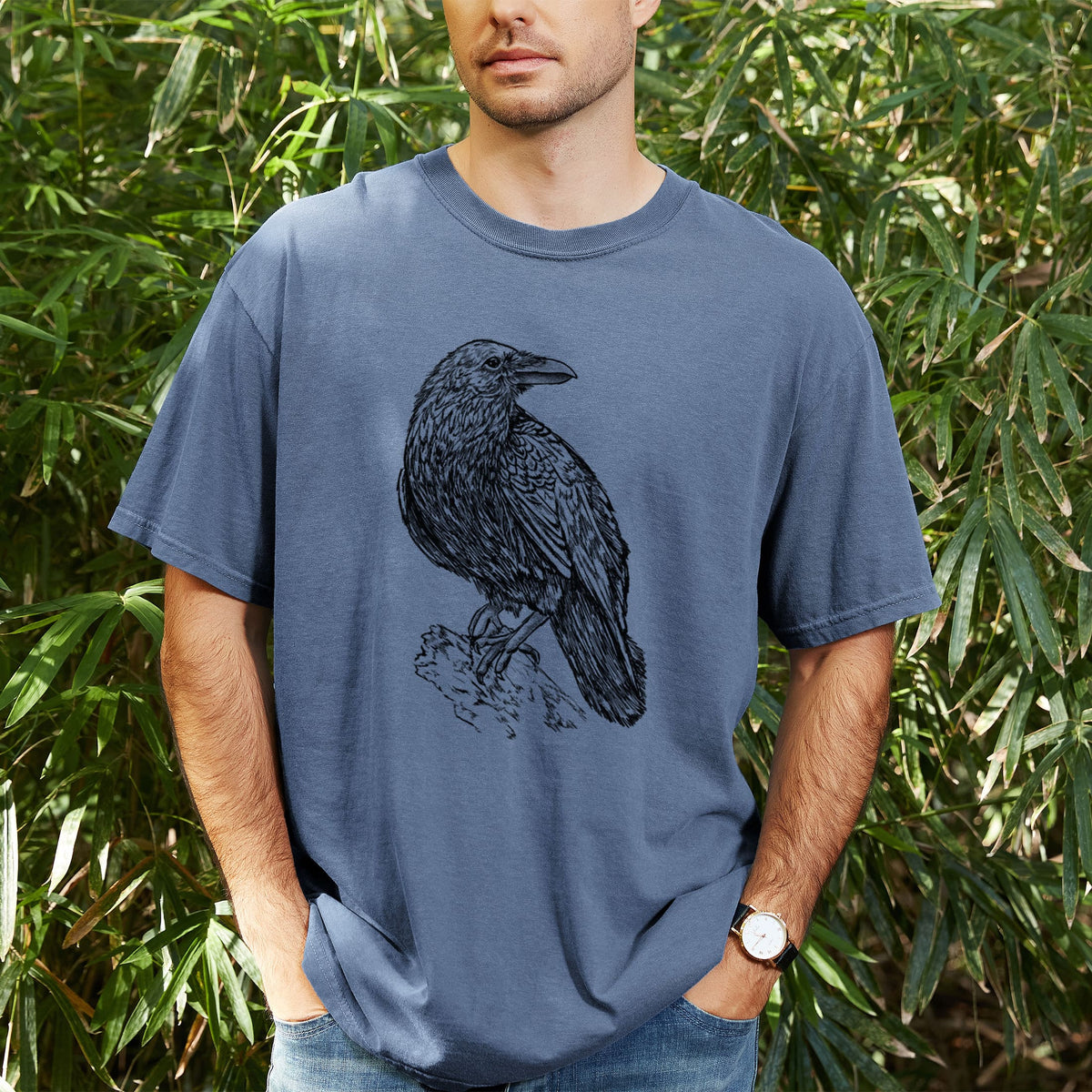 Man wearing a Comfort Colors t-shirt printed with a detailed black ink sketch of a common raven perched on a rock, outdoors.