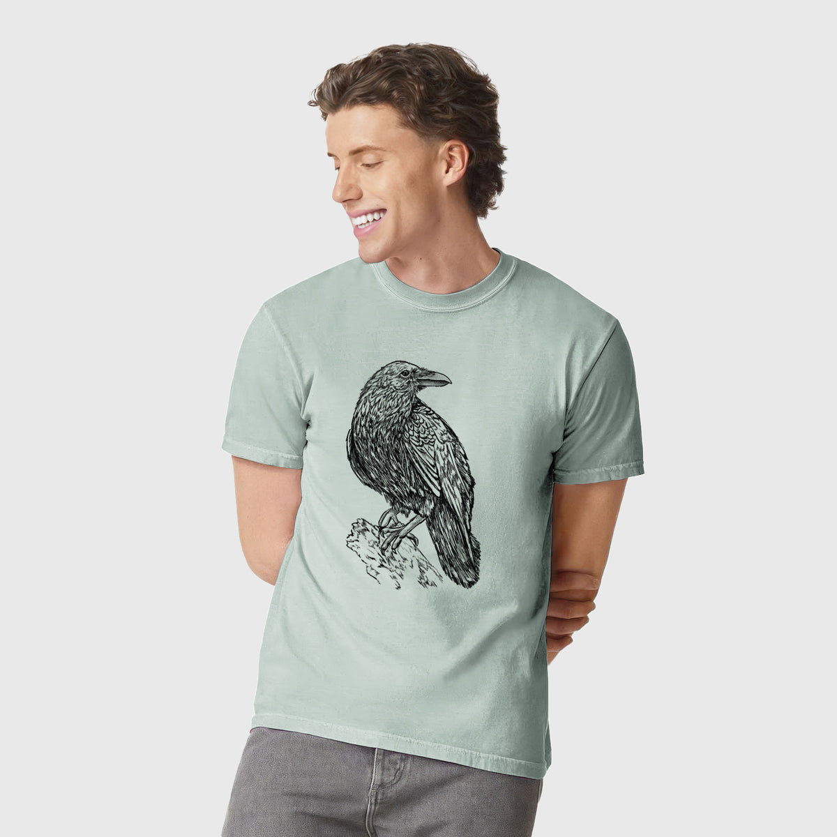 Man wearing a Comfort Colors t-shirt printed with a detailed black ink sketch of a common raven perched on a rock.