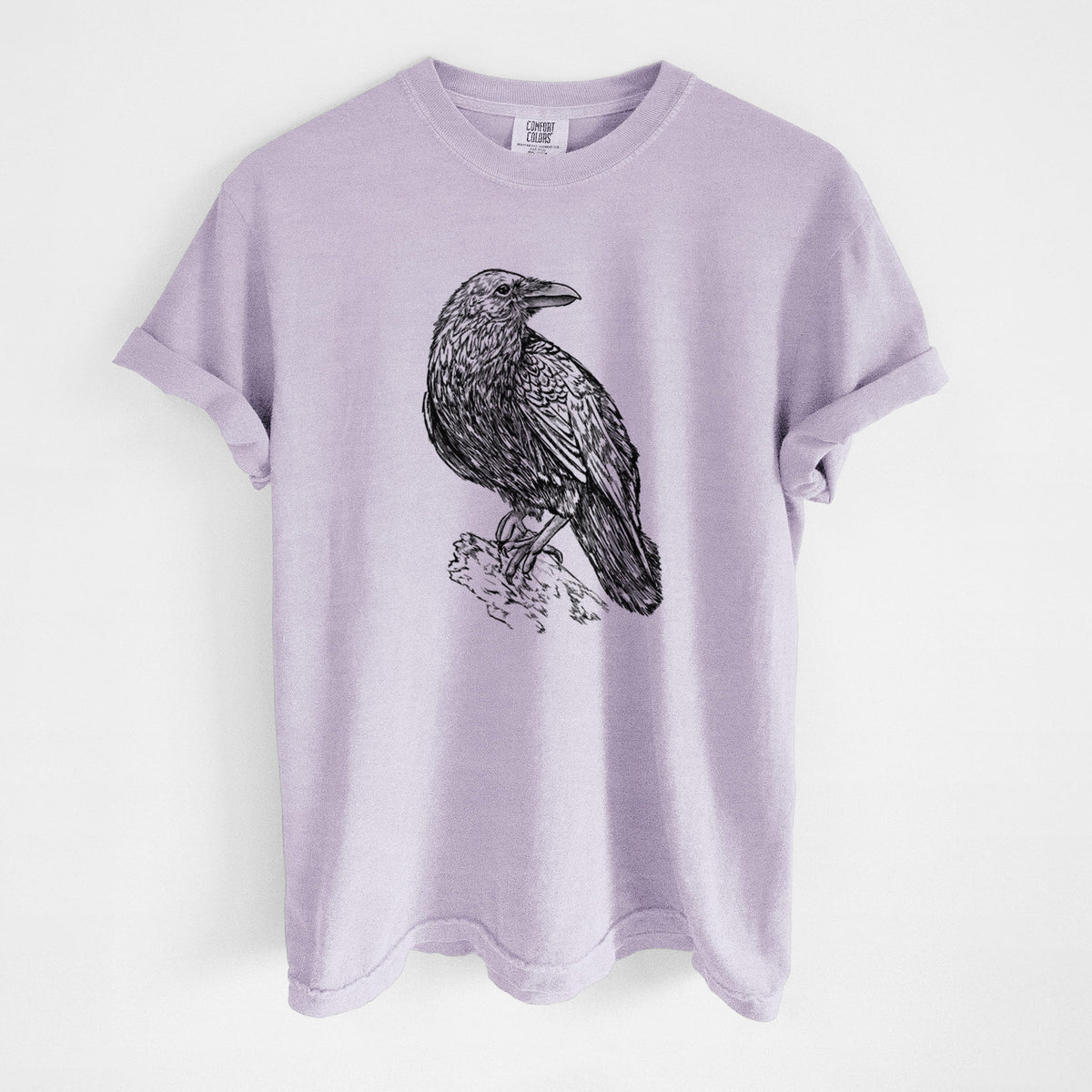 Orchid purple Comfort Colors t-shirt laid flat, printed with a detailed black ink sketch of a common raven perched on a rock.