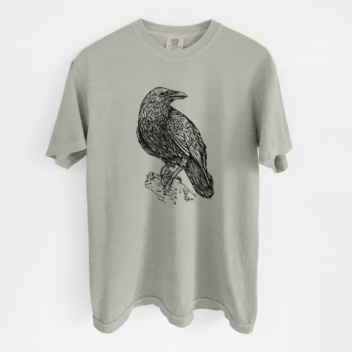 Sandstone beige Comfort Colors t-shirt laid flat, printed with a detailed black ink sketch of a common raven perched on a rock.
