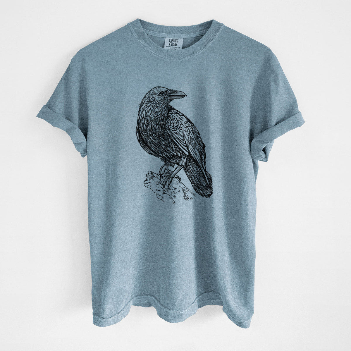 Steel blue Comfort Colors t-shirt laid flat, printed with a detailed black ink sketch of a common raven perched on a rock.