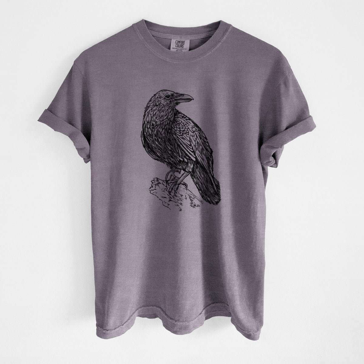Wine red Comfort Colors t-shirt laid flat, printed with a detailed black ink sketch of a common raven perched on a rock.