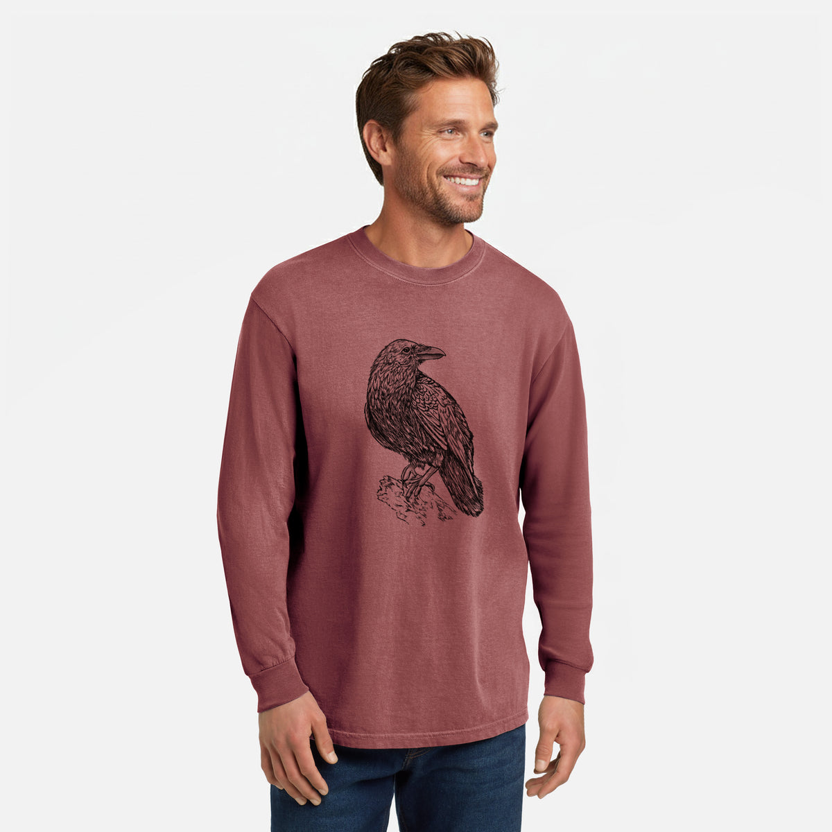 Brick red Comfort Colors long sleeve shirt printed with a detailed black ink sketch of a common raven perched on a rock, shown in a lifestyle photo.