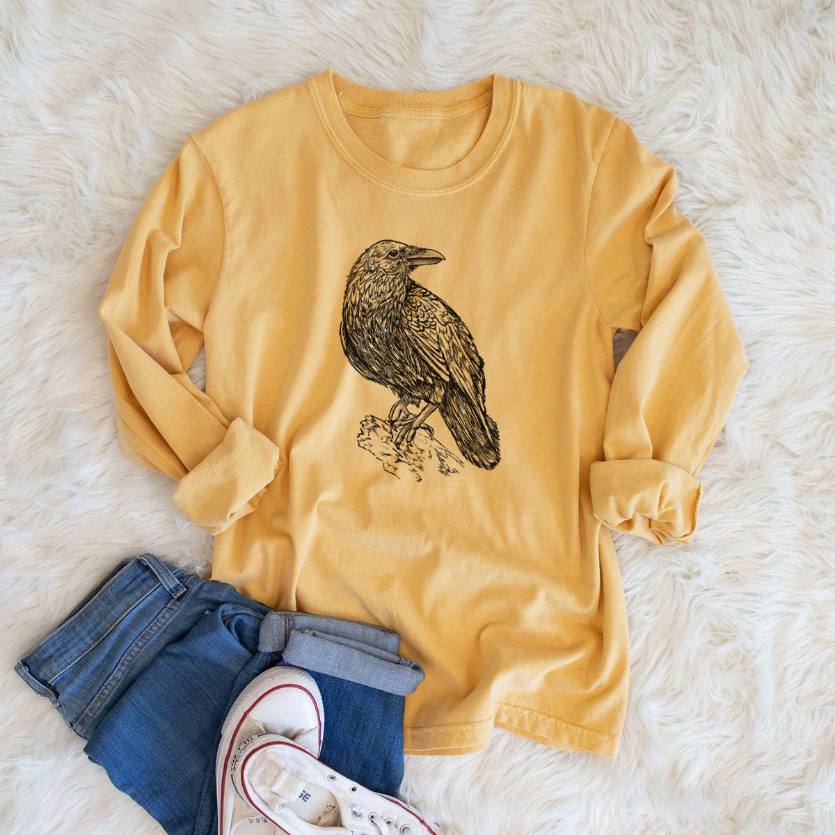 Golden wheat yellow Comfort Colors long sleeve shirt printed with a detailed black ink sketch of a common raven perched on a rock, shown in a lifestyle photo.