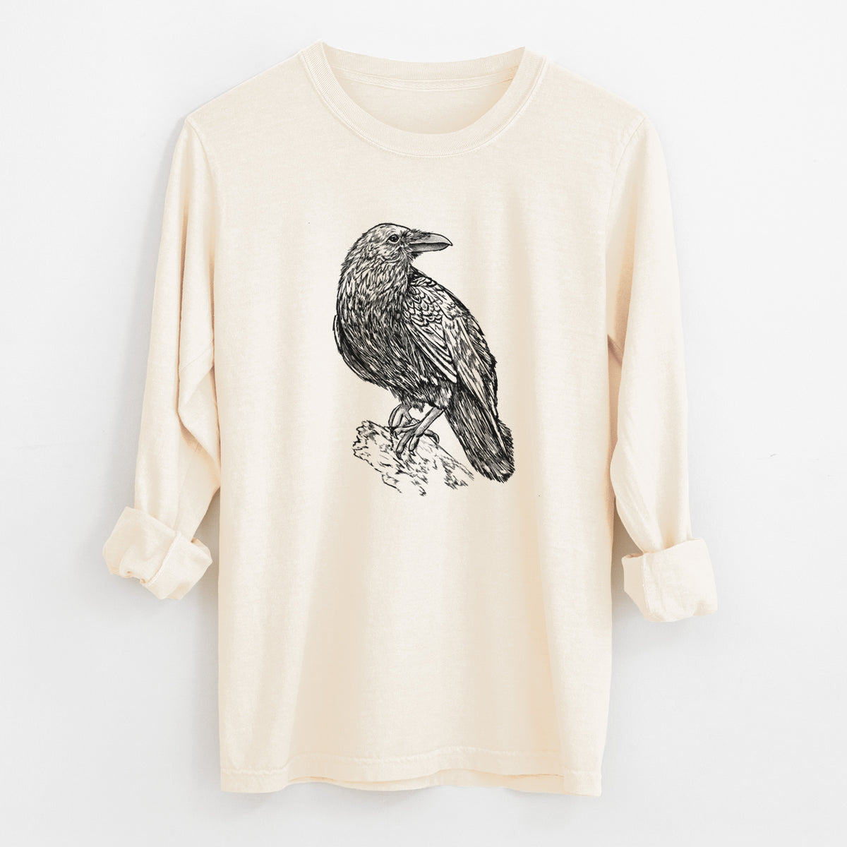 Ivory Comfort Colors long sleeve shirt laid flat with the cuffs rolled, printed with a detailed black ink sketch of a common raven perched on a rock.