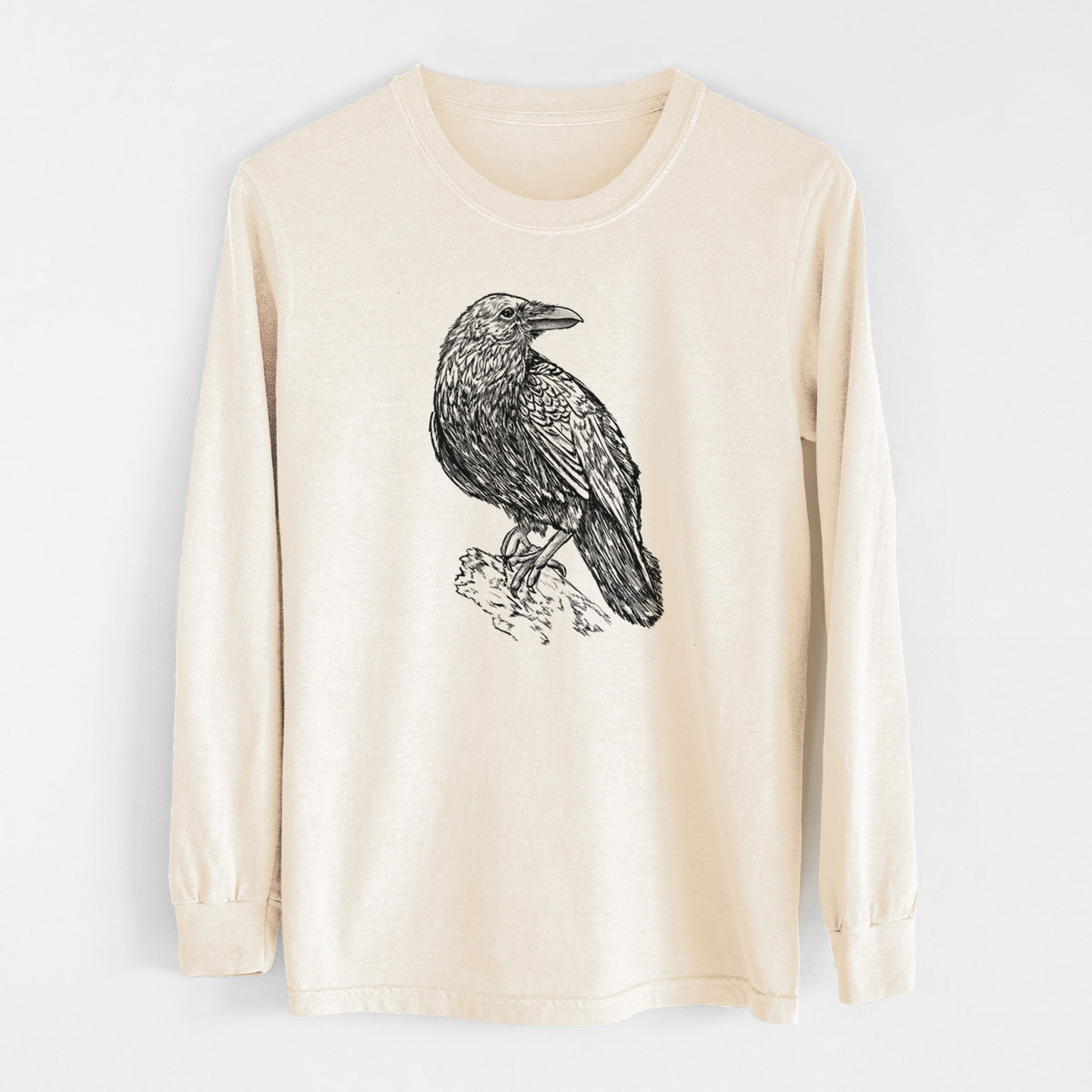 Ivory Comfort Colors long sleeve shirt laid flat, printed with a detailed black ink sketch of a common raven perched on a rock.